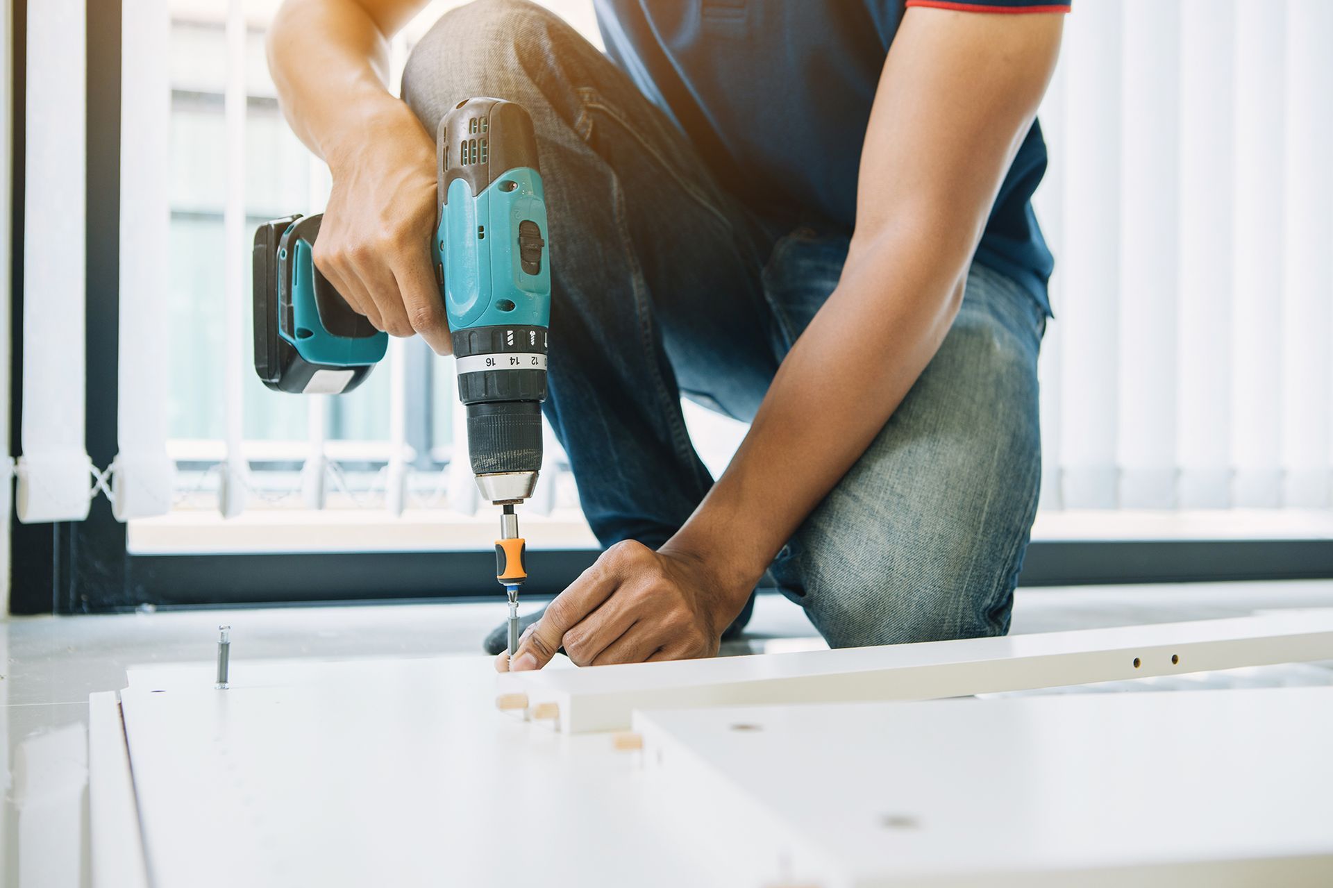 A man is kneeling down while using a drill on a piece of wood.