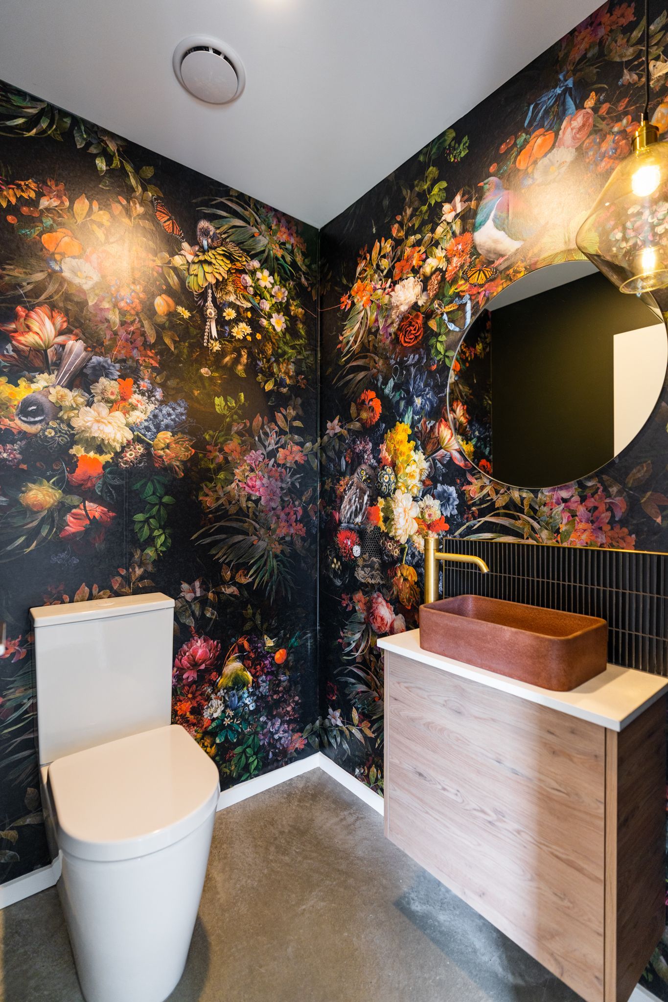 A bathroom with a toilet , sink , mirror and floral wallpaper.