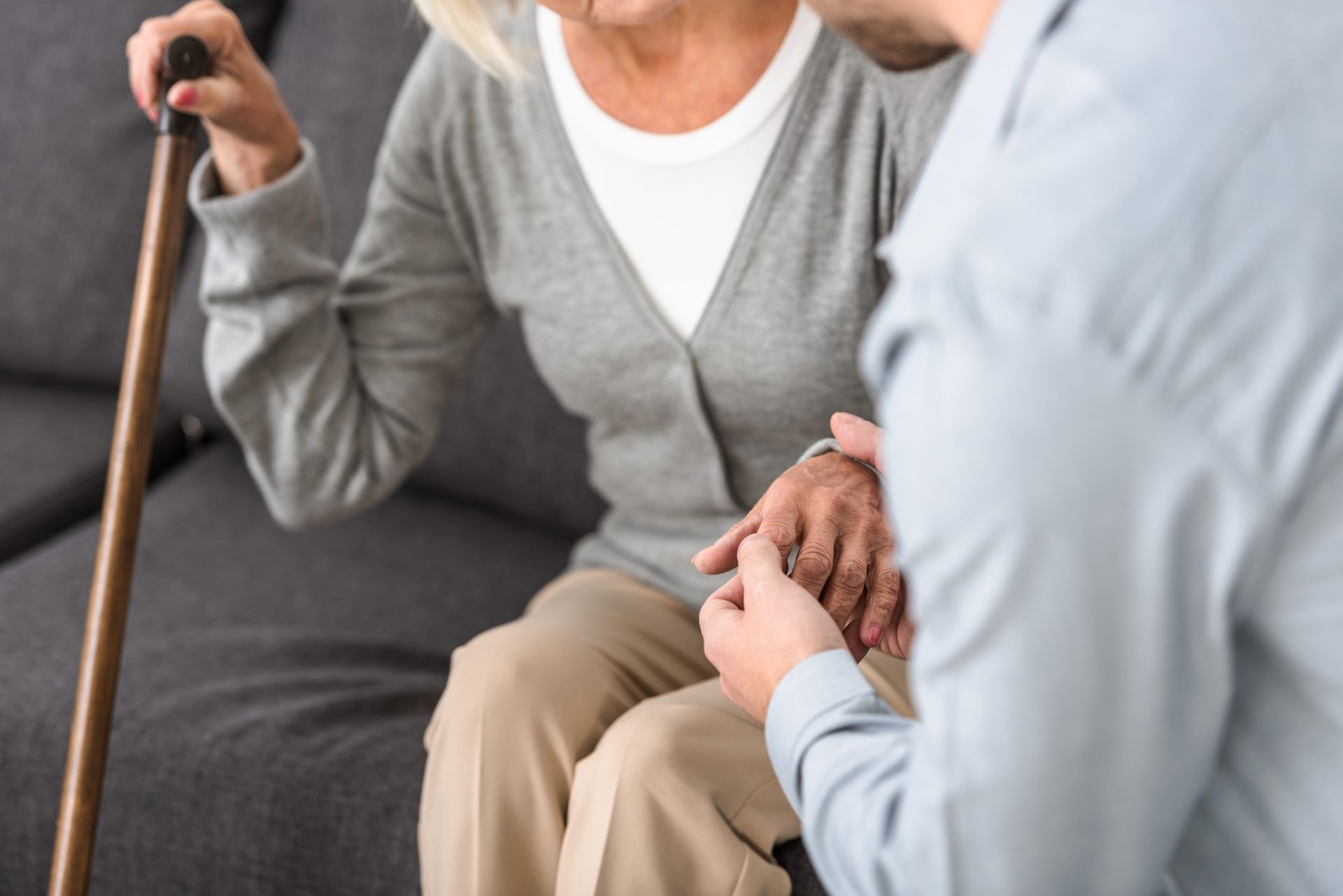 A person holds the hand of an individual sitting on a sofa and holding a walking cane.
