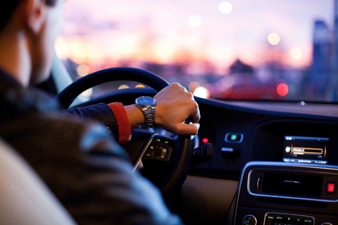 Person driving a car at dusk, holding the steering wheel. City lights in background.