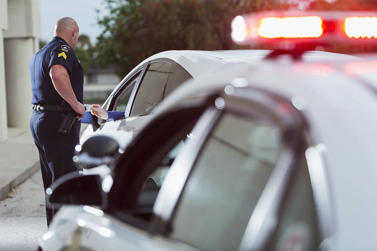 Police officer issuing a ticket to a driver; red and white police lights are on.