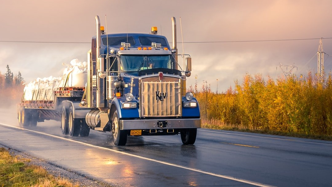 Blue semi-truck driving on a wet road, hauling cargo; overcast sky and yellow foliage in the background.