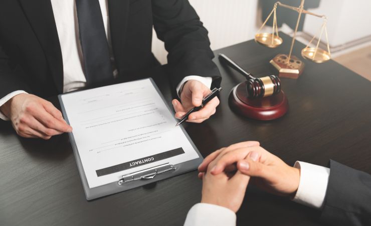 Lawyer pointing pen at contract with client, gavel, and scales of justice on desk.