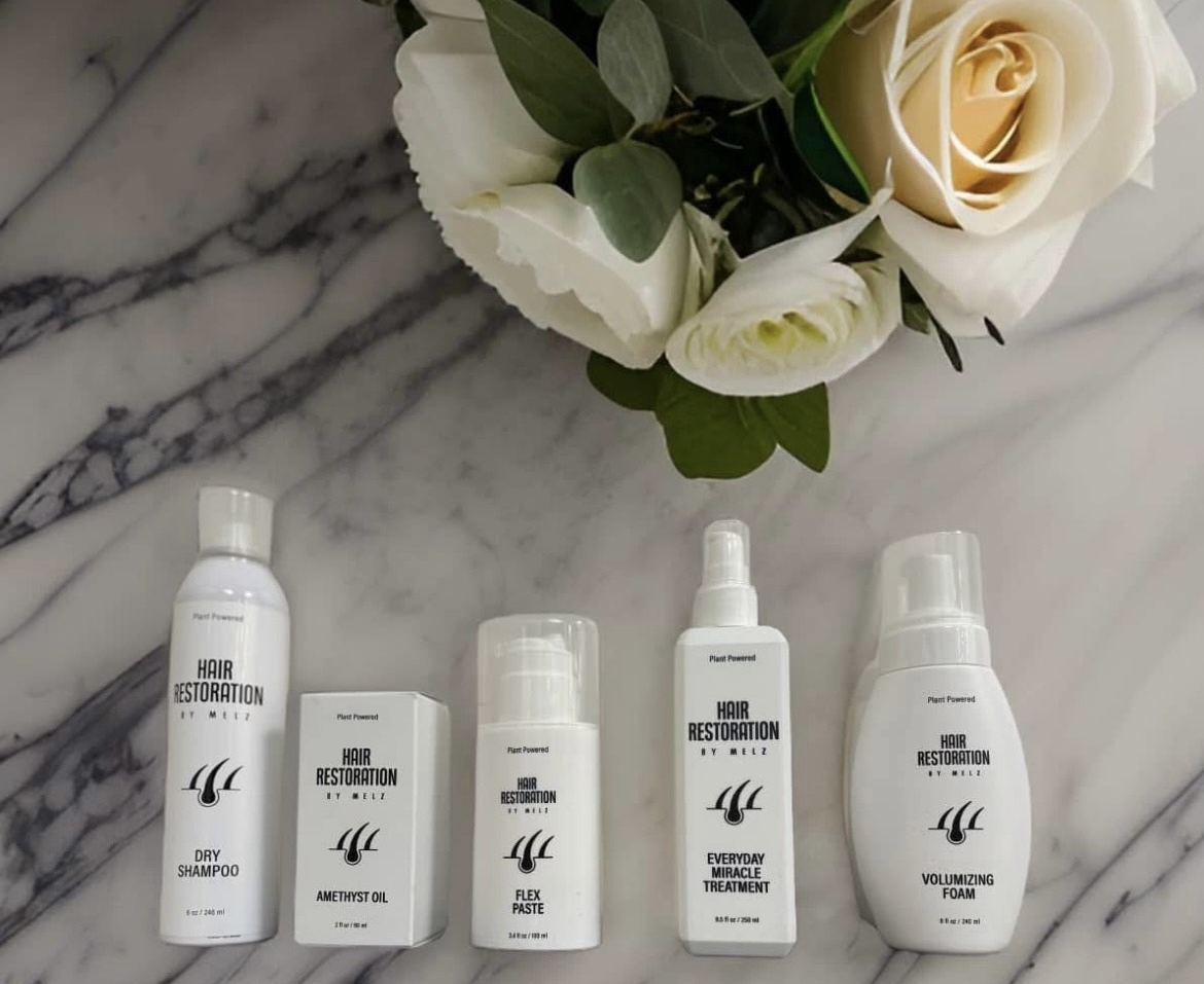 Five white hair care products, arranged below a bouquet of white flowers on a marble surface.
