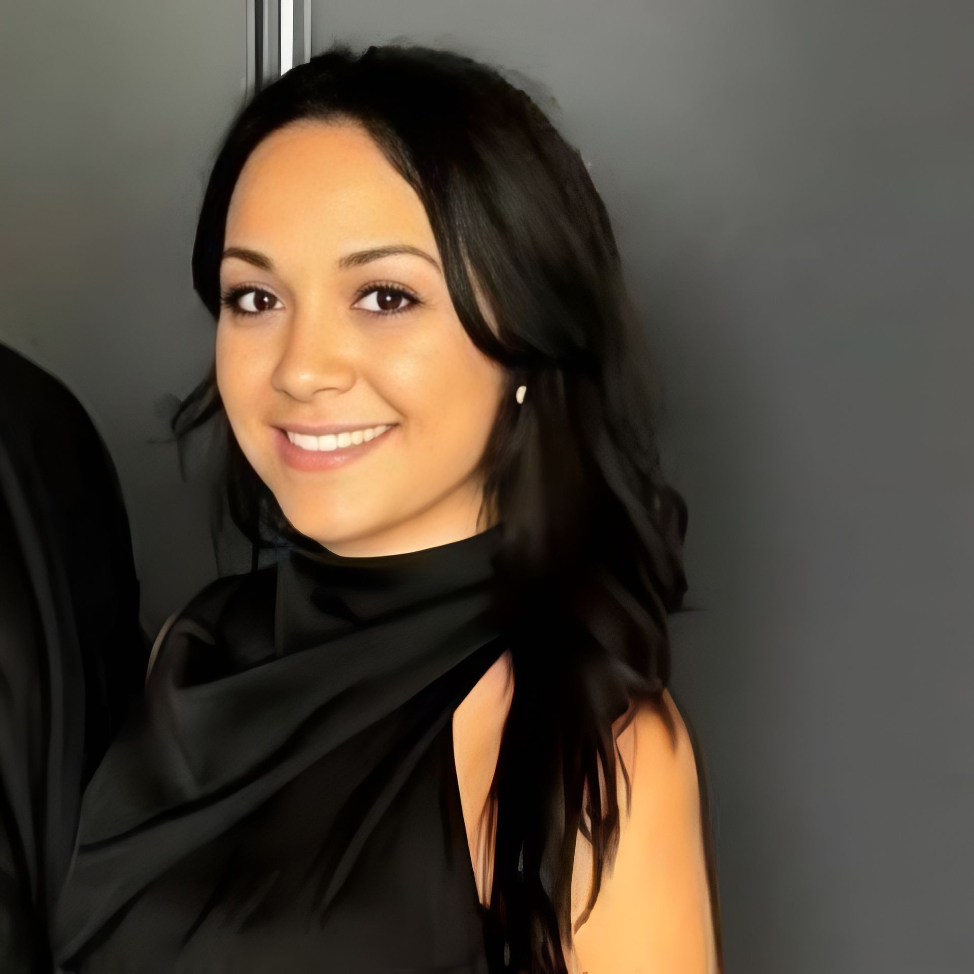Woman with dark hair smiles, wearing a black dress, and posing against a gray background.