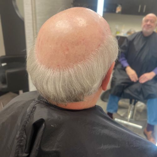 A view from behind of an individual in a salon cape with a horseshoe-shaped hairstyle, reflected in a mirror.