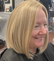 A person with blonde, shoulder-length bob hair smiling while sitting in a salon chair.