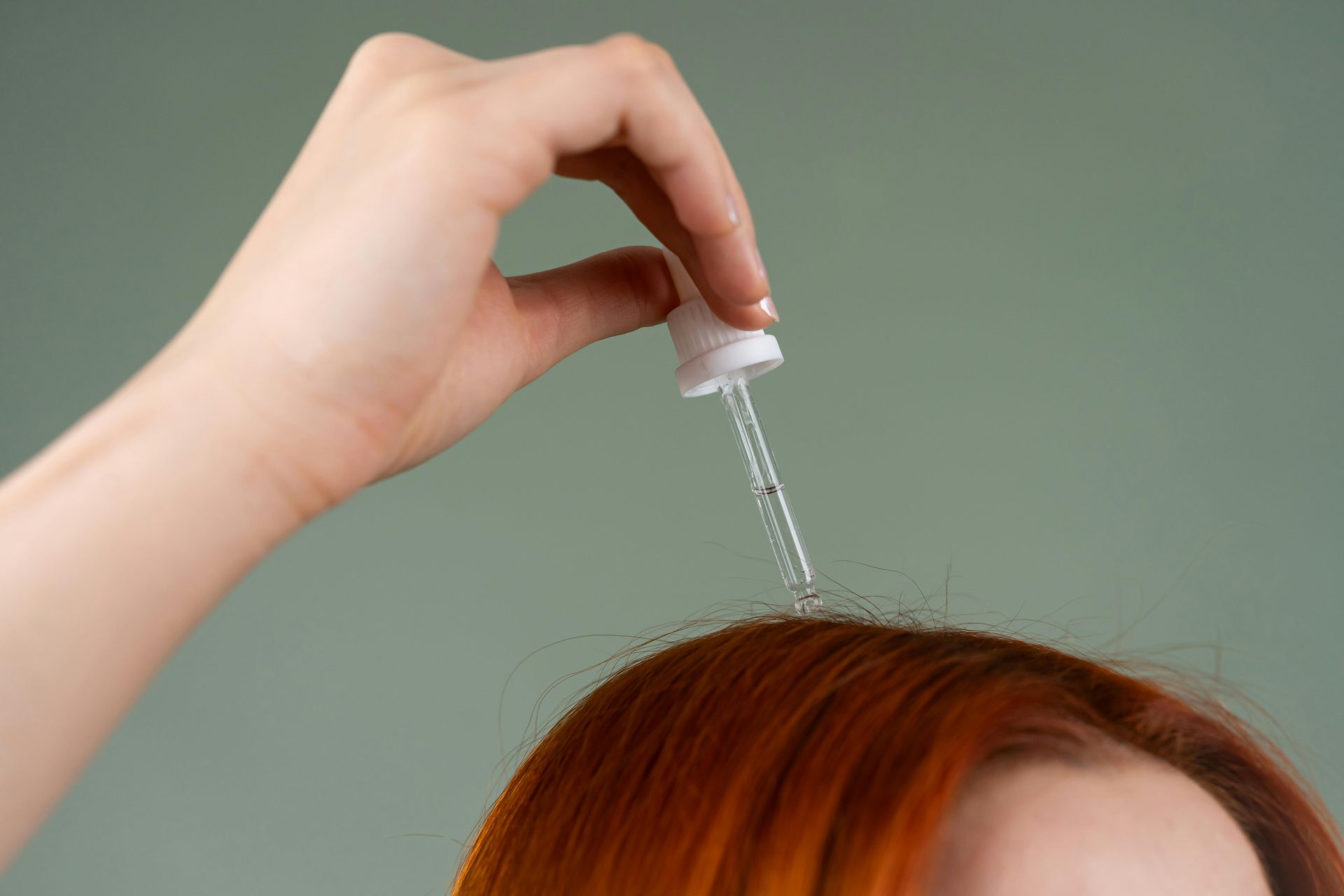 Hand holding dropper, applying liquid to red hair on a green background.
