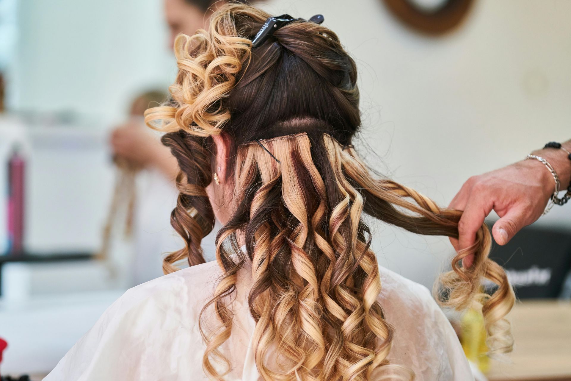 Woman with curly hair extensions being styled in a salon. A stylist is holding a strand.