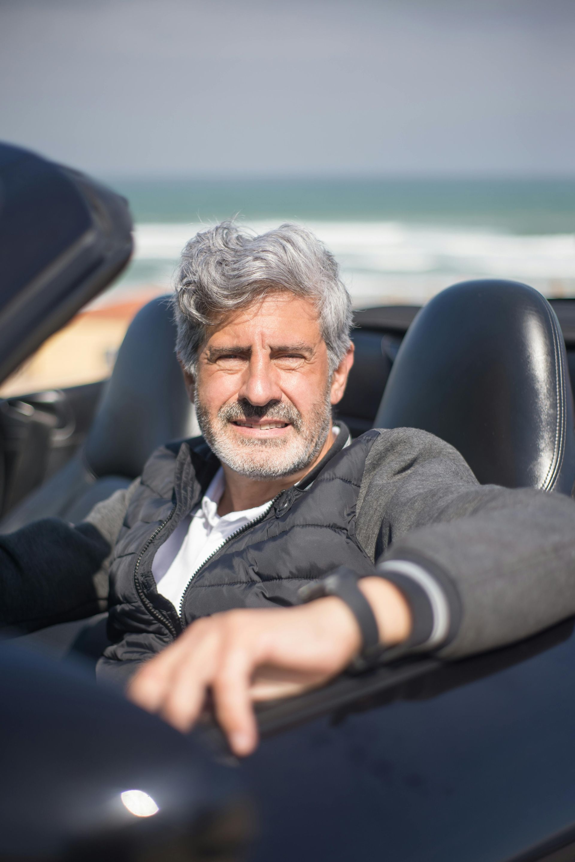 A person with gray hair and a beard smiling while sitting in a convertible car with the ocean in the background.