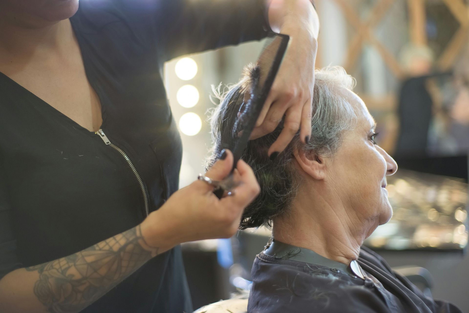 Hairdresser cuts an older woman's hair with scissors and comb in a salon, smiling.
