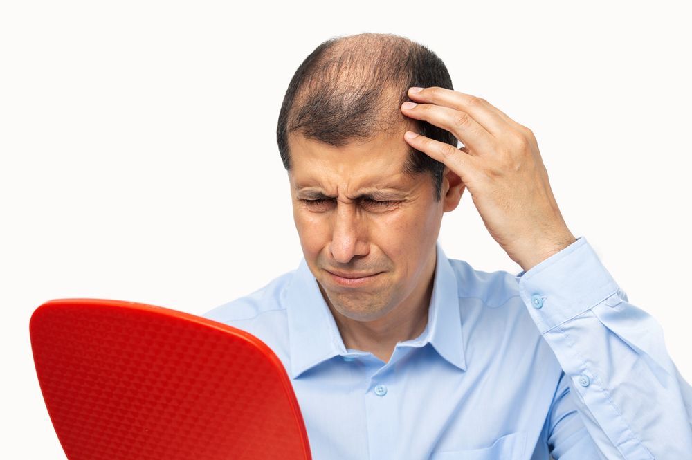 Man with thinning hair looks distressed at his reflection in a red hand mirror.