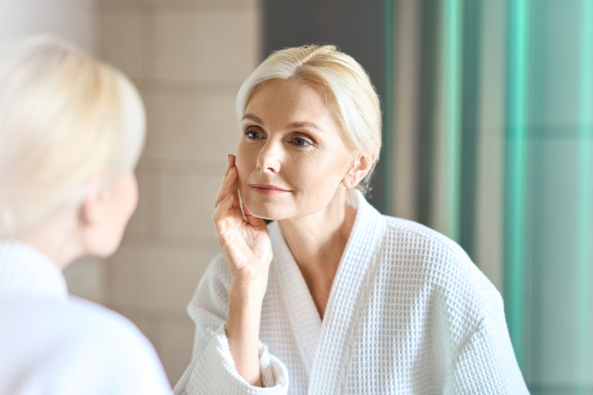 Blonde woman in a white robe examines her face in a mirror, lit by natural light.