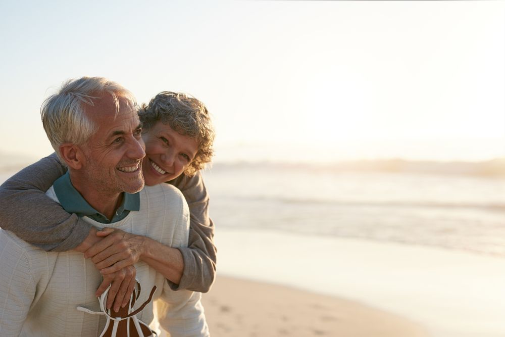 Older couple embracing on beach, woman with arms around man, both smiling.
