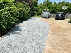 Gravel driveway next to a concrete driveway, two cars parked near a basketball hoop.