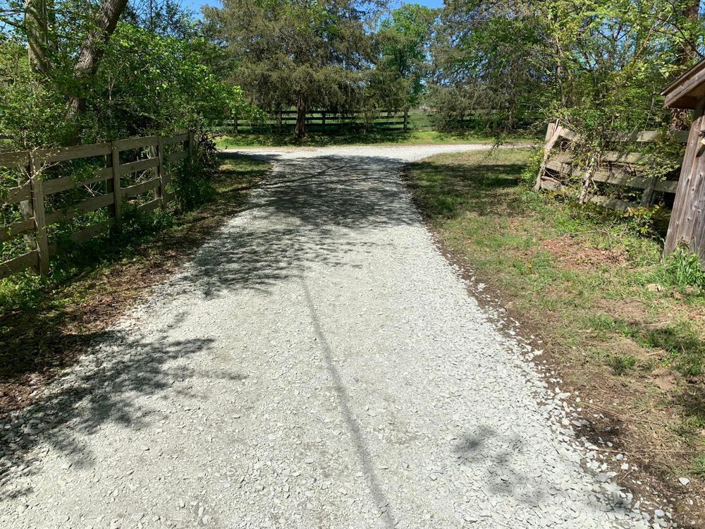 Gravel driveway splits into two paths, with wooden fences and trees on either side, outdoors on a sunny day.