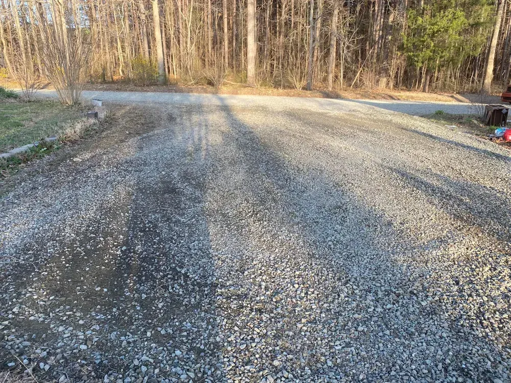 Gravel driveway in sunlight, leading to trees; a small patch of darker, possibly wet, gravel near the entrance.