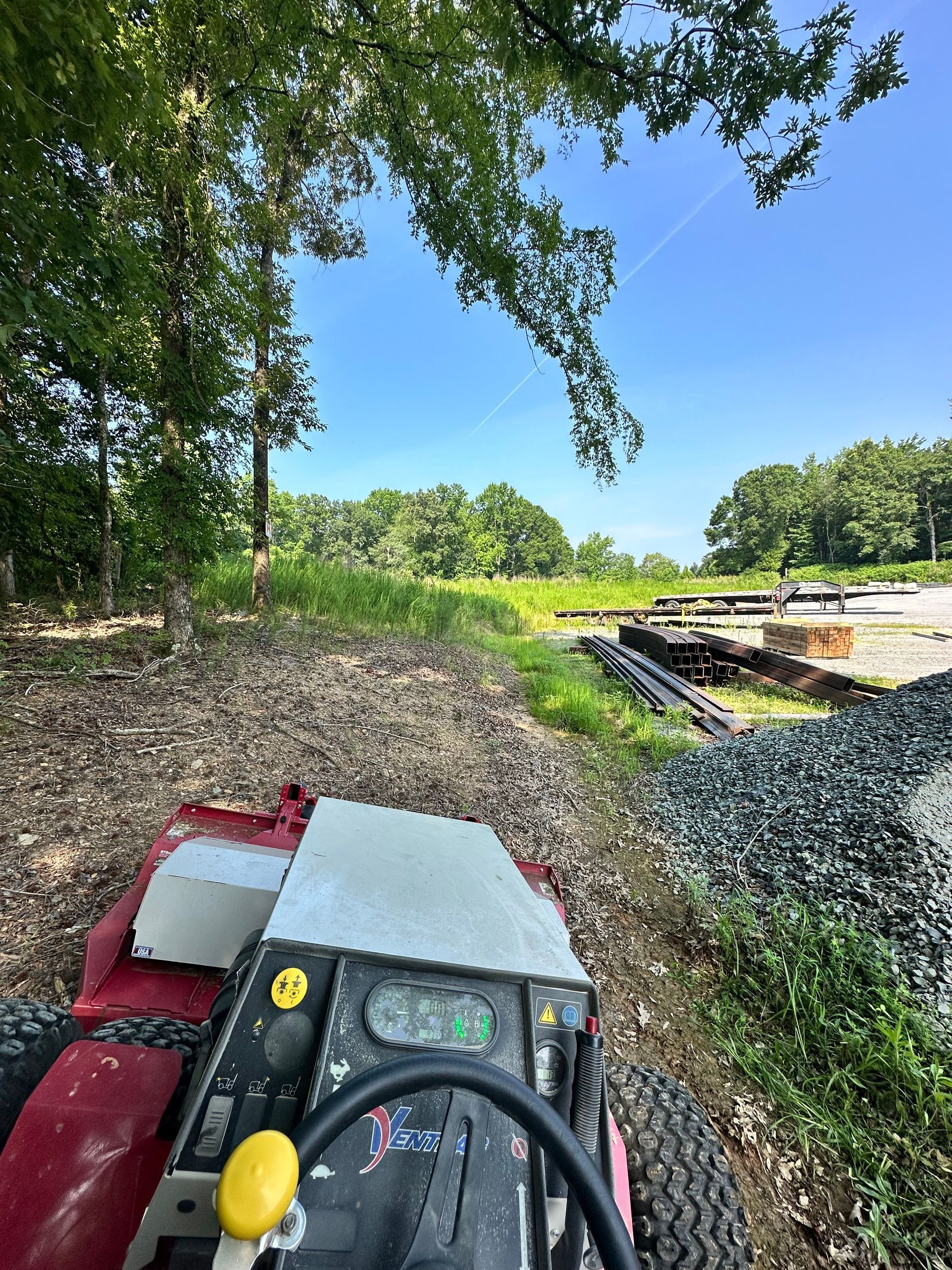 Red tractor mowing overgrown grass in a sunny field, trees on both sides, blue sky.