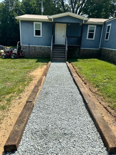 Gravel pathway bordered by wooden beams leads to a blue house with a porch and stairs.
