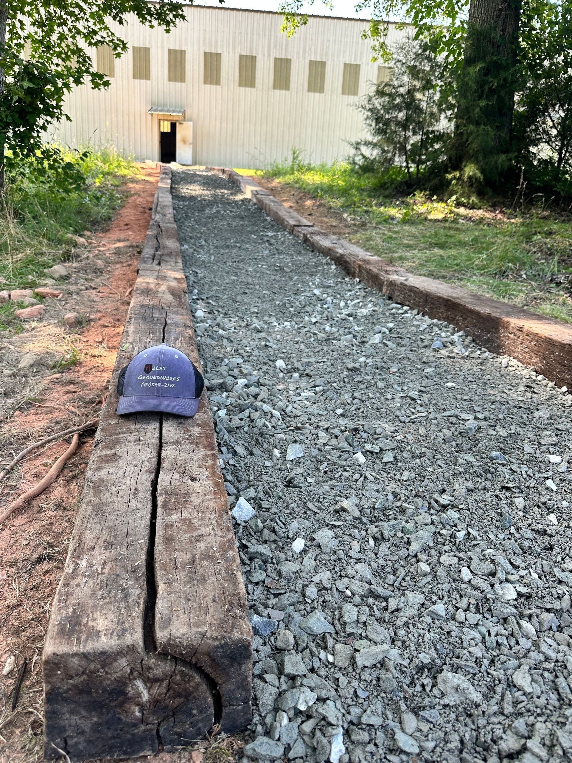 Gravel path bordered by wooden beams leading to a building. Blue hat rests on a beam.