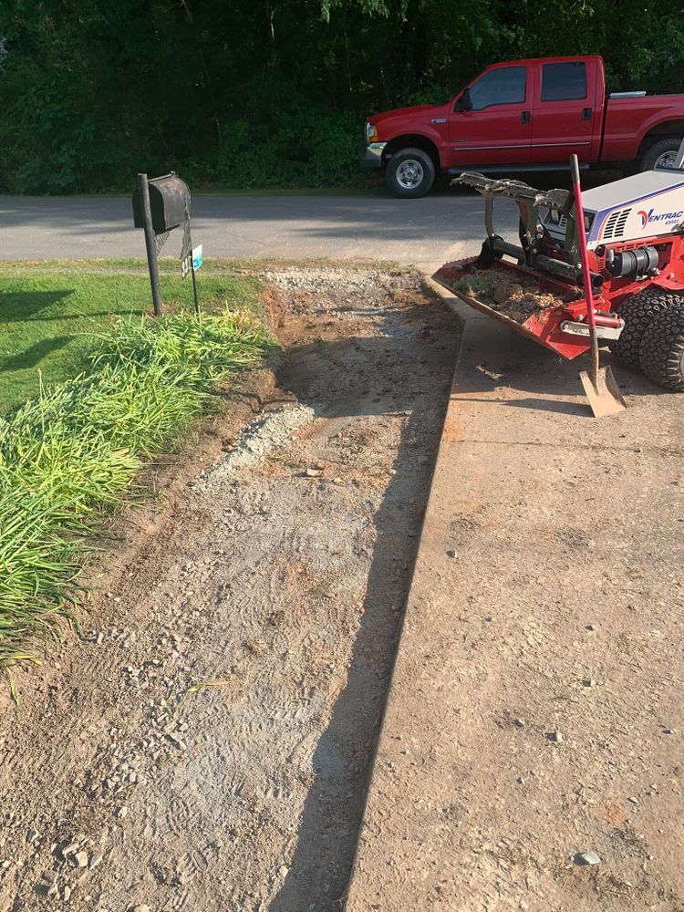 A trencher cuts a line in a gravel driveway, next to a red truck and mailbox on a sunny day.