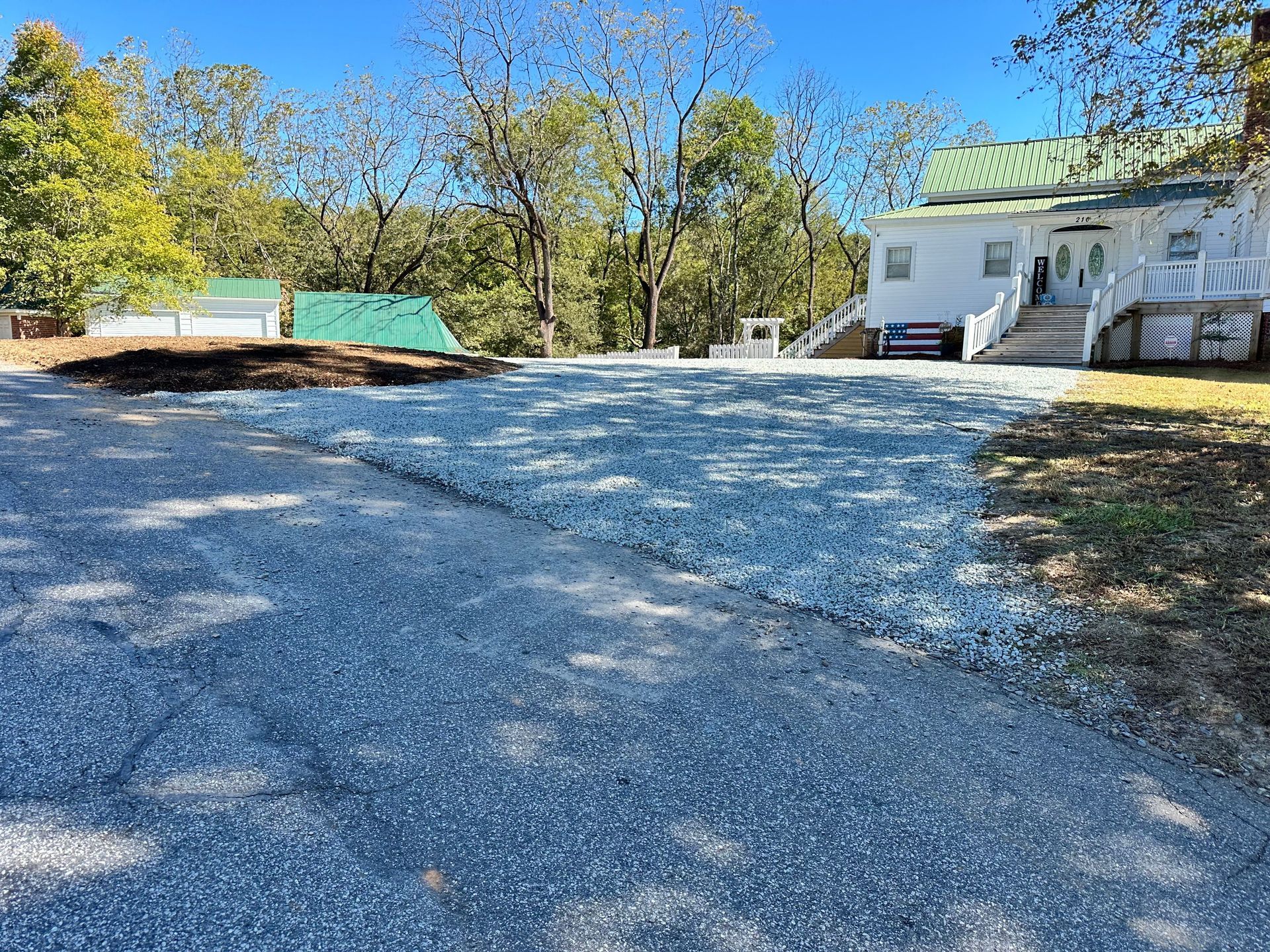 Gravel driveway in front of a white house on a sunny day. Green tarp and trees in the background.