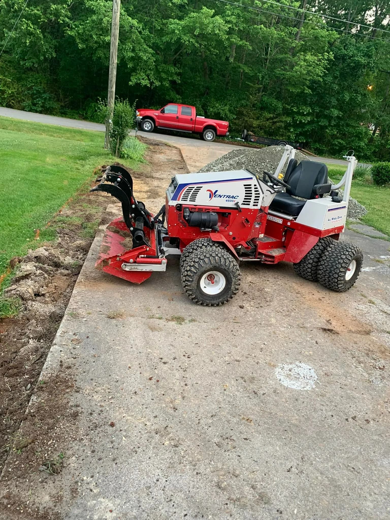 Red and white snowblower on a concrete path, red pickup truck in the background, grassy area and trees.