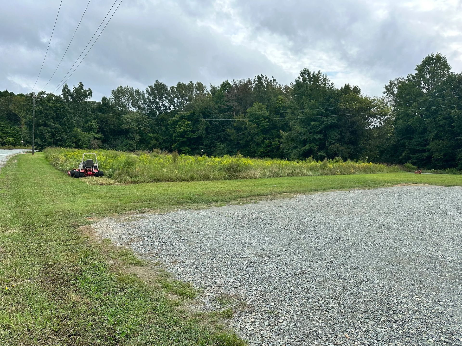 Gravel parking area beside a grassy field; a tractor mows grass near the woods under an overcast sky.