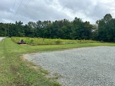 Gravel parking area beside a grassy field; a tractor mows grass near the woods under an overcast sky.