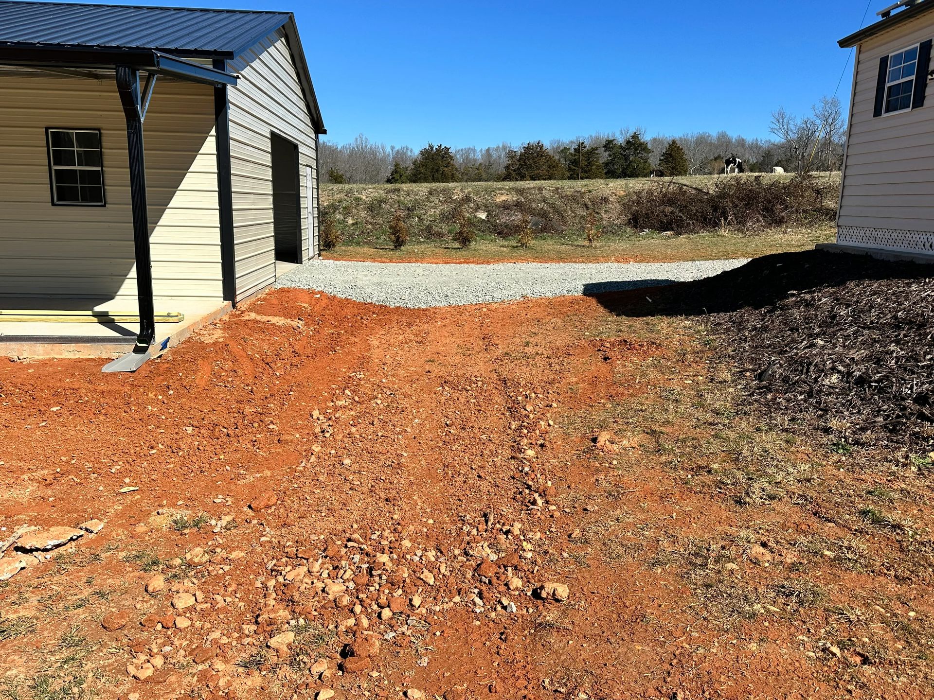 Red dirt path leading to a gravel driveway and a light-colored shed on a sunny day.