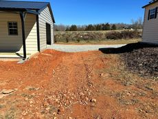 Red dirt path leading to a gravel driveway and a light-colored shed on a sunny day.