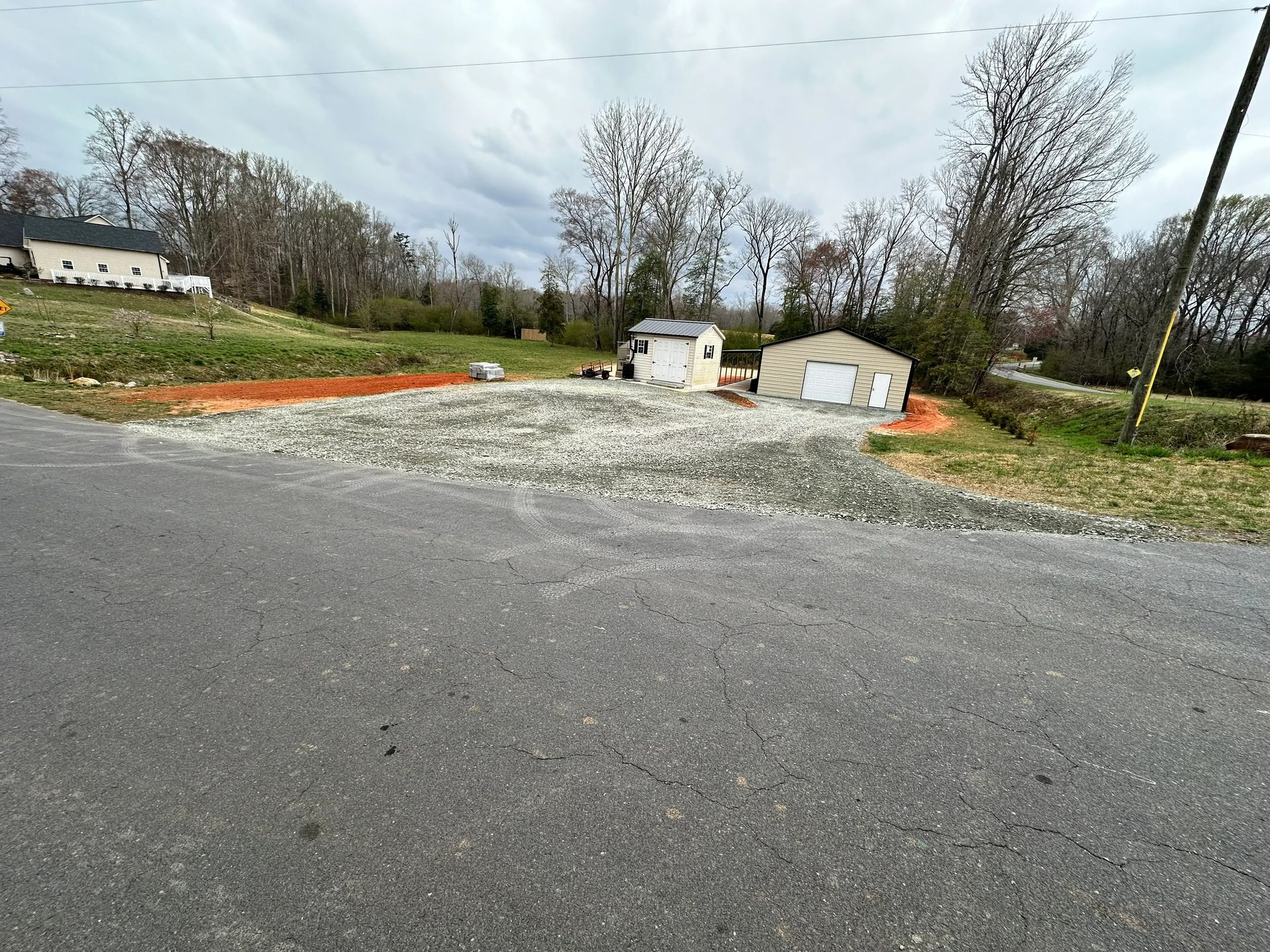 Gravel driveway with two small buildings, one with a garage door. A house is on a hill in the background.