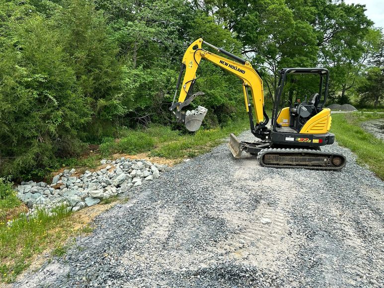 Yellow excavator working on a gravel pile near a green tree line.