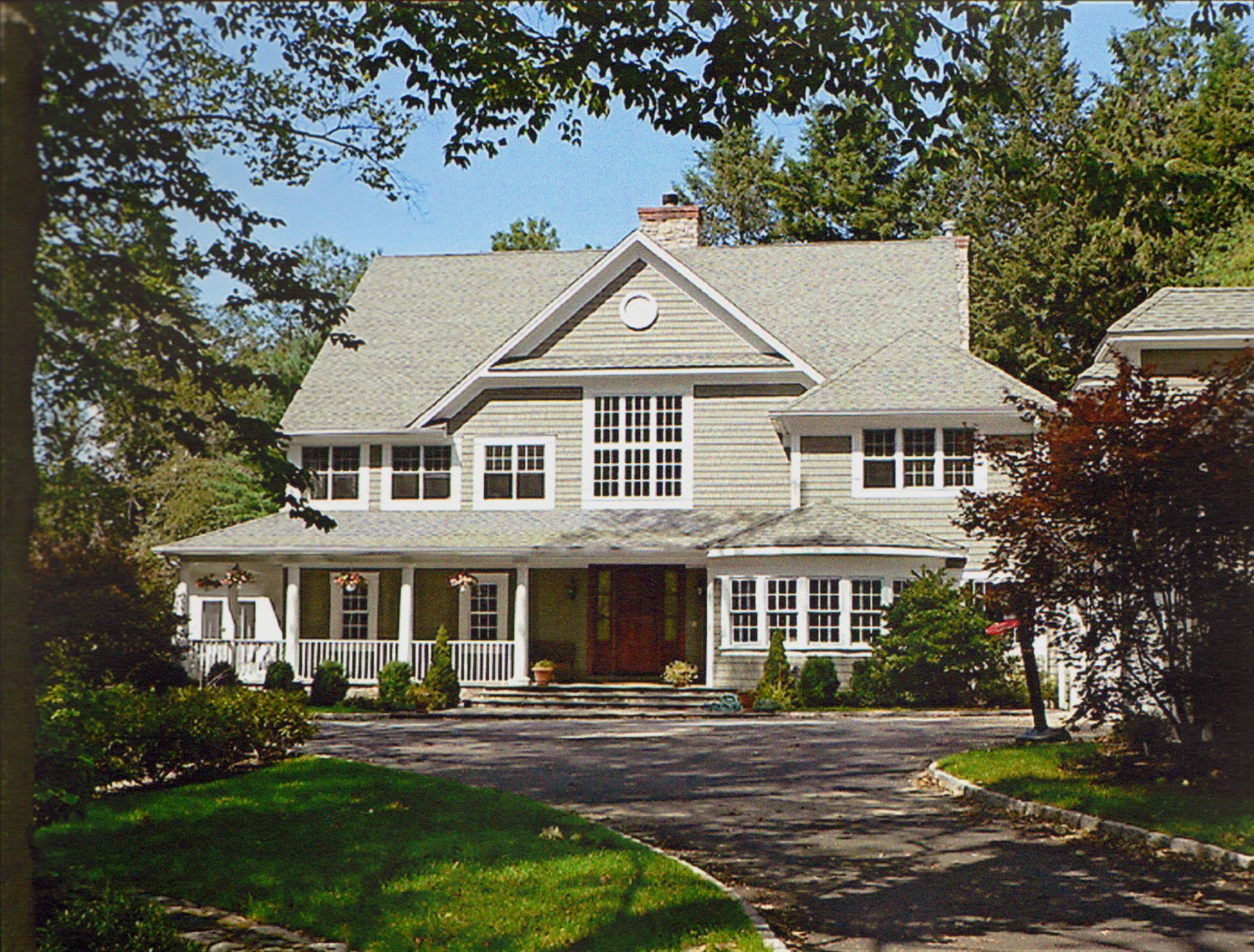 Two-story house with light gray siding, white trim, and a porch. Green lawn and trees surround the driveway.