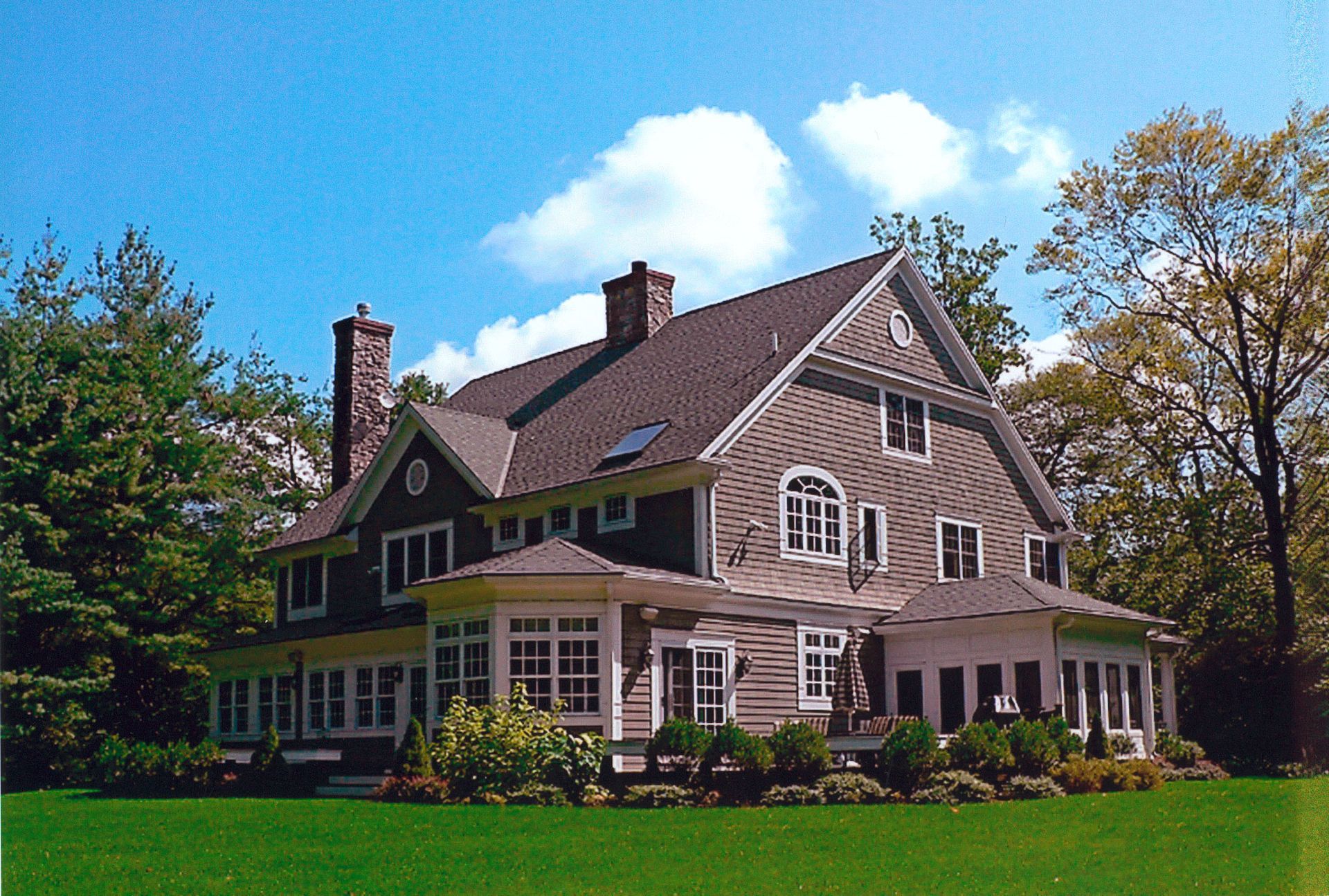 Large, multi-story house with gray siding, two chimneys, and wraparound porch, on a green lawn under a blue sky.