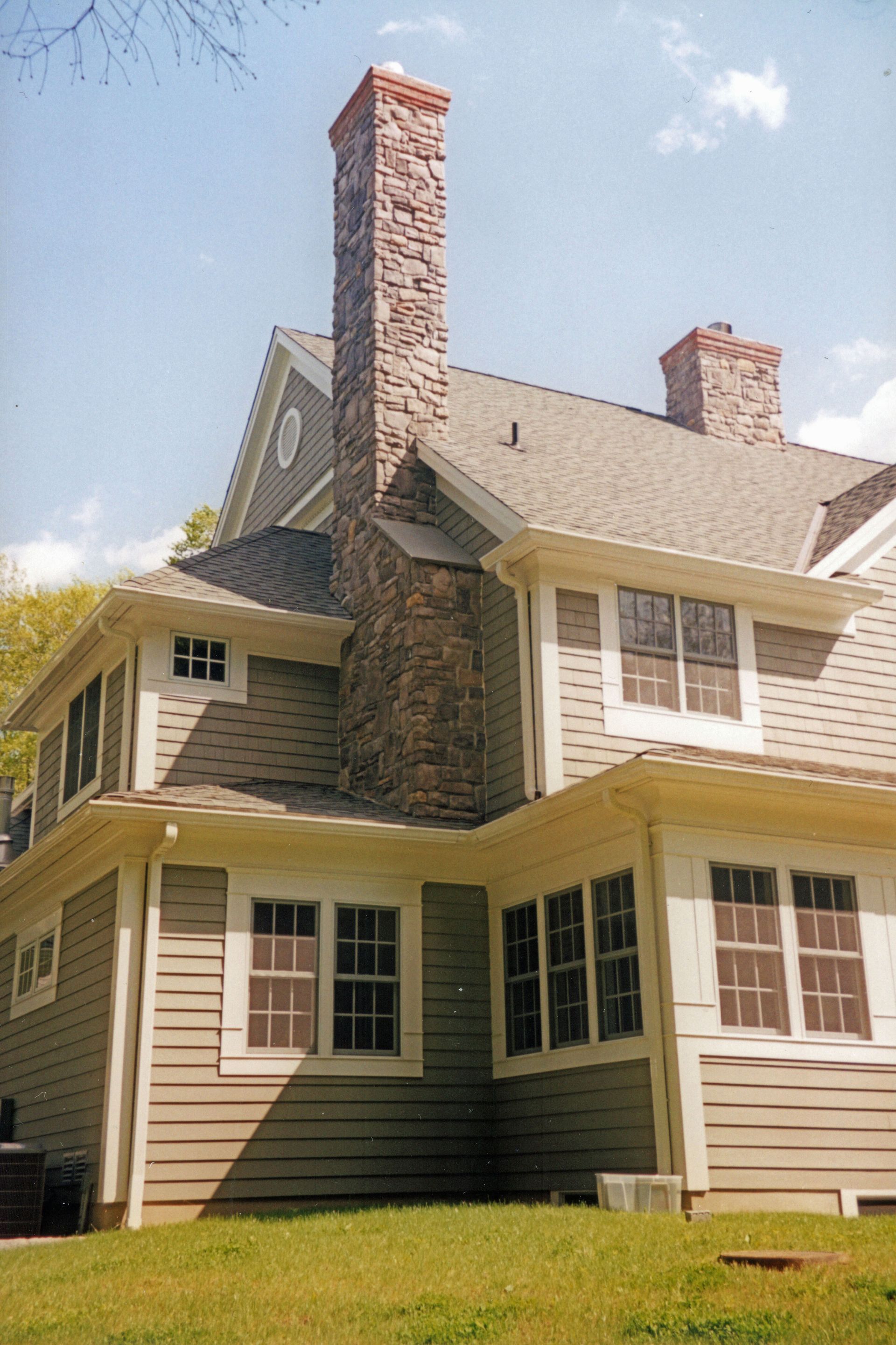 Stone chimney on a two-story house with tan siding, several windows, and a gray shingled roof.