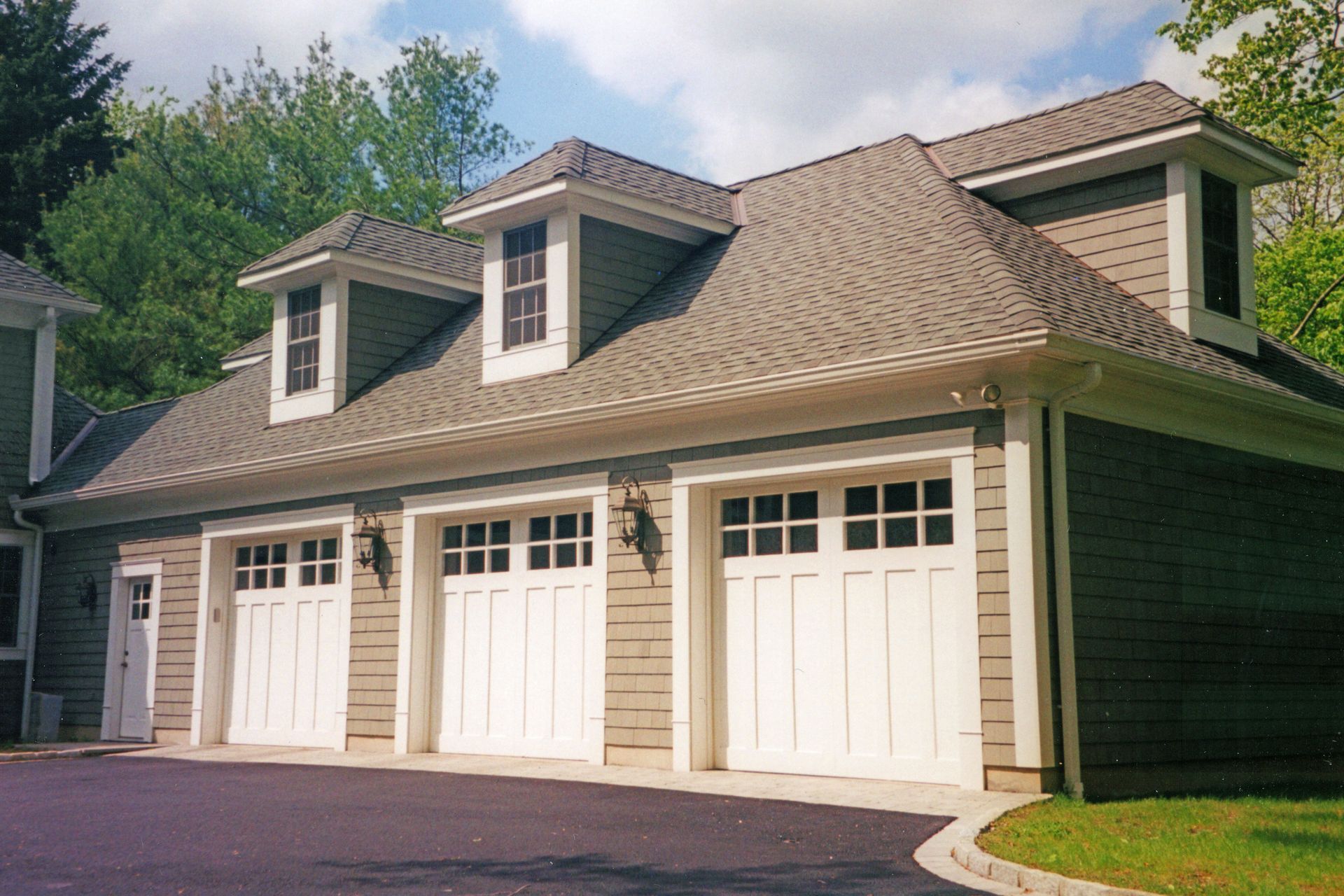 Three-car garage with white doors, tan siding, and dormers under a gray shingled roof.
