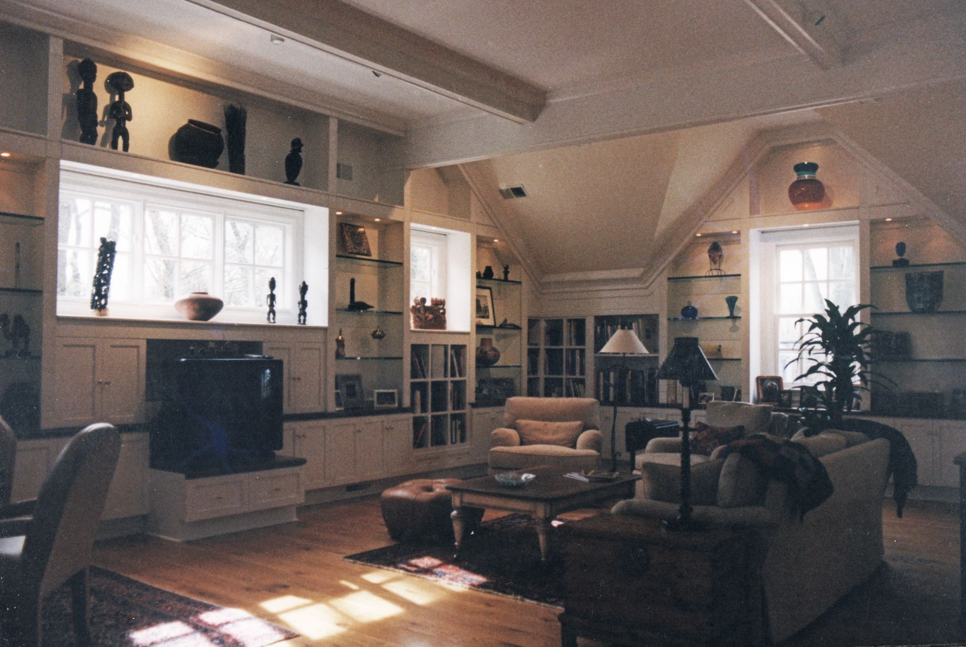 Living room with built-in shelves and display cabinets, displaying artwork; two armchairs, a sofa, and windows.