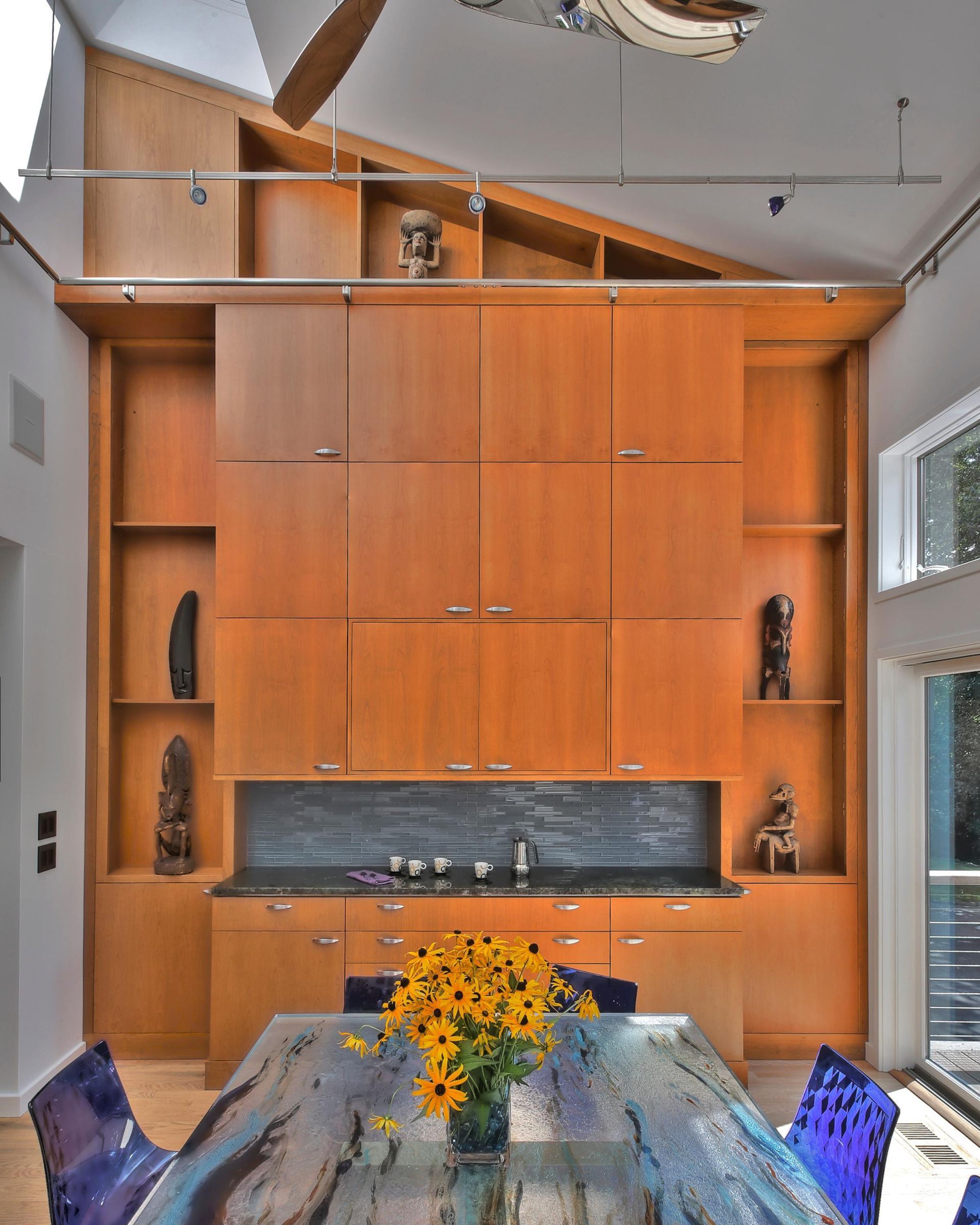 Wooden built-in with cabinets, shelves, and a small sink, dining table with yellow flowers, and blue chairs.