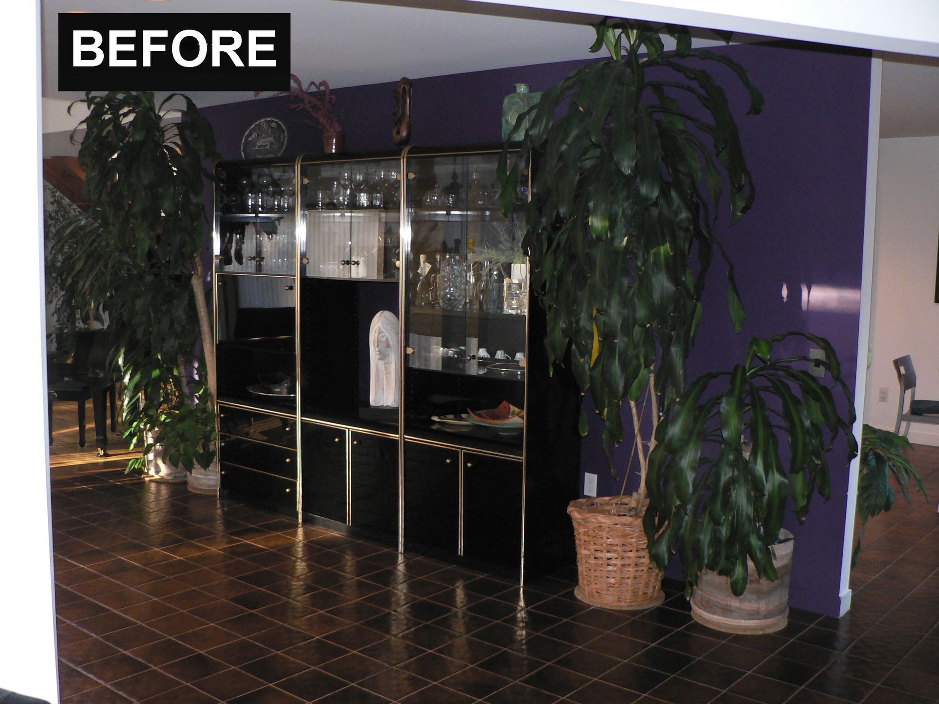 A dark wood-floored room with a black and gold cabinet and purple wall. Large plants flank the cabinet.