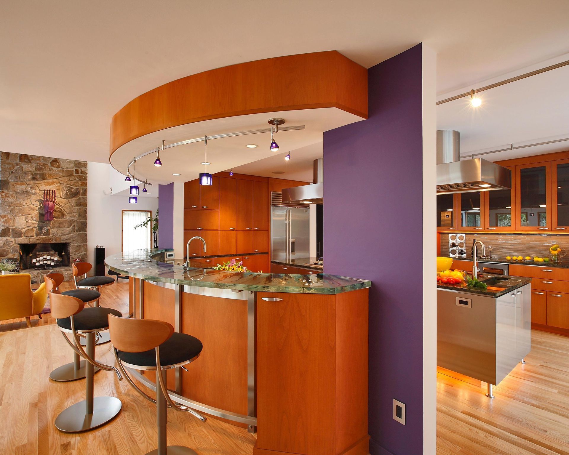 Modern kitchen with curved wooden bar, purple accent wall, stainless steel appliances, and wooden cabinetry.