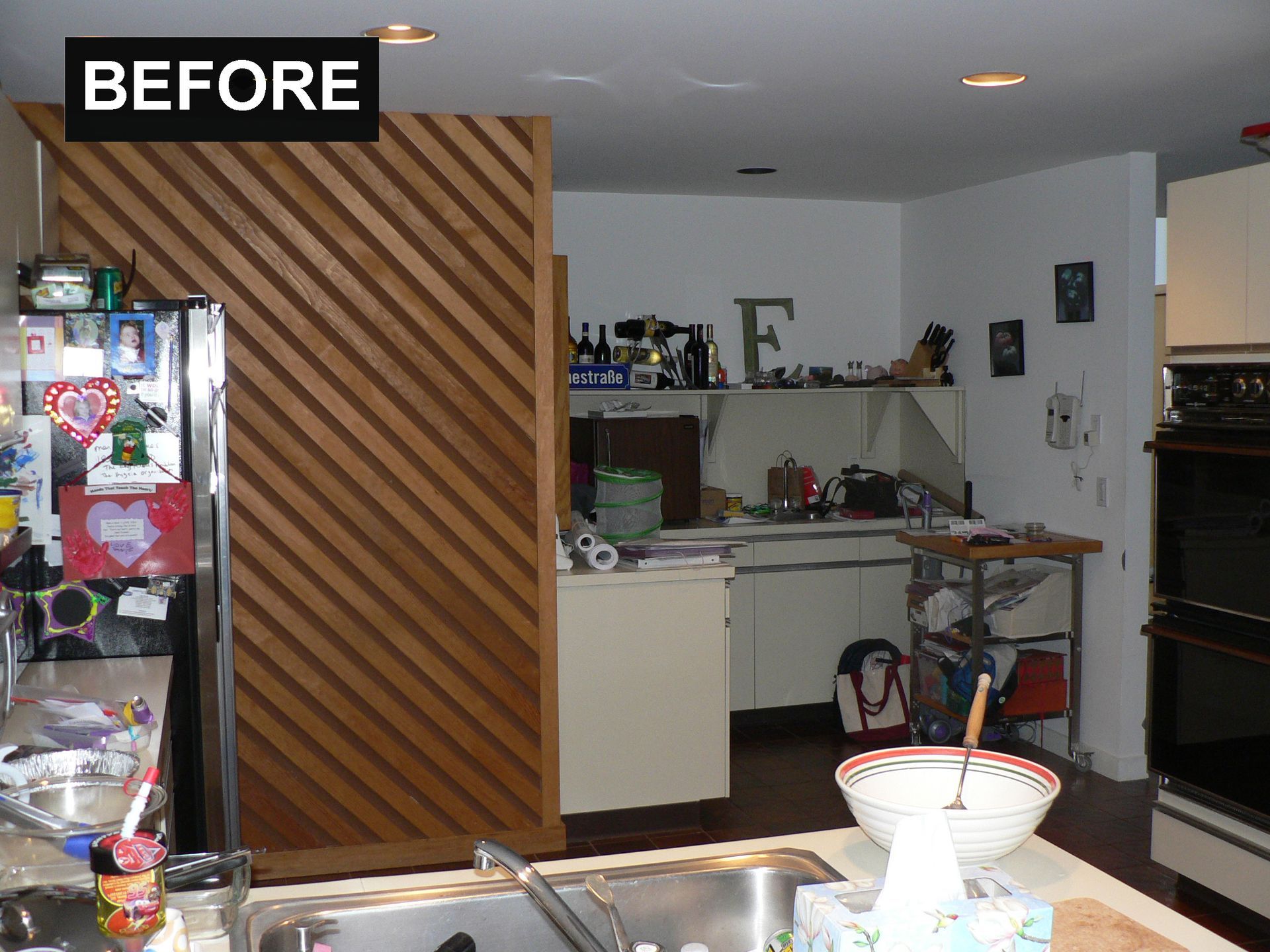 Kitchen before renovation; cluttered countertops, diagonal wooden divider, stainless steel appliances.