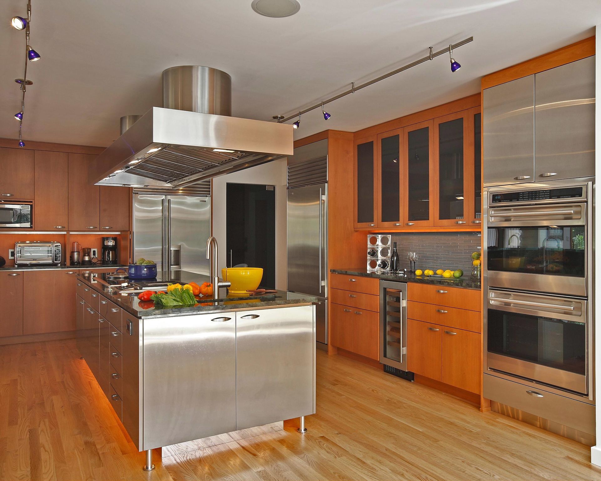 Modern kitchen with stainless steel island, cabinets, and appliances; wood floor and track lighting.