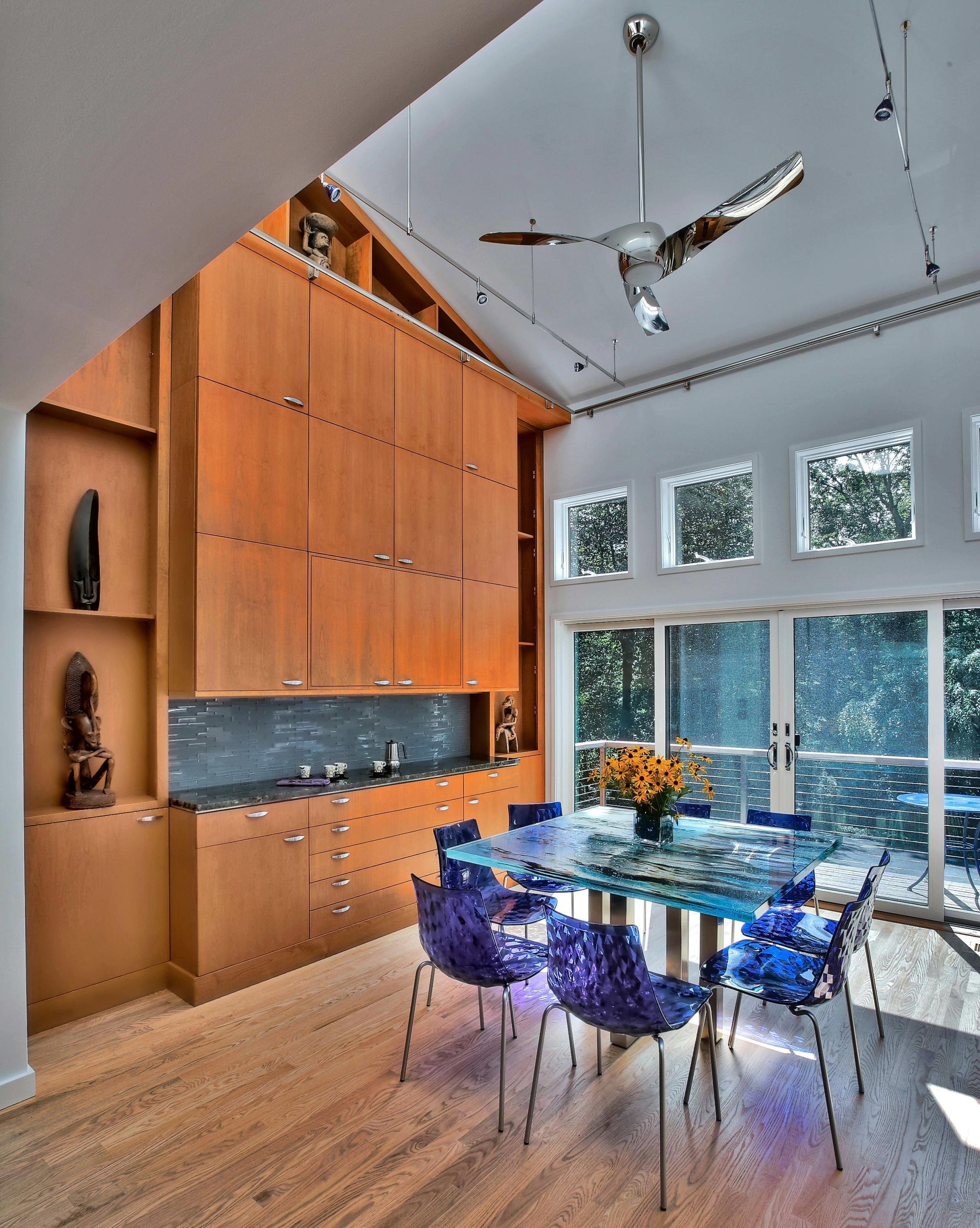 Modern dining room with glass table, blue chairs, and wood cabinetry. High ceilings and large windows.