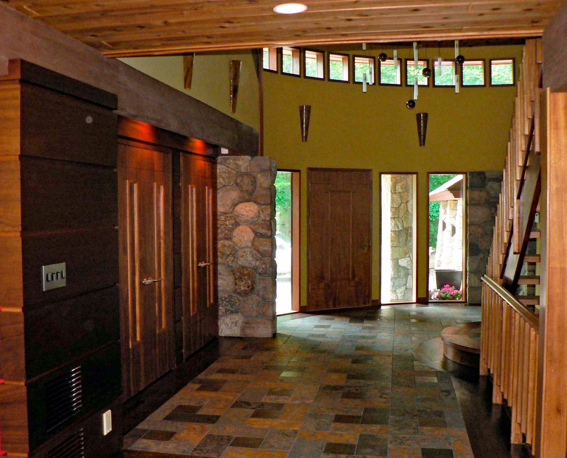 Interior view of a hallway with wood and stone features; dark cabinets on left, entrance with large windows ahead.