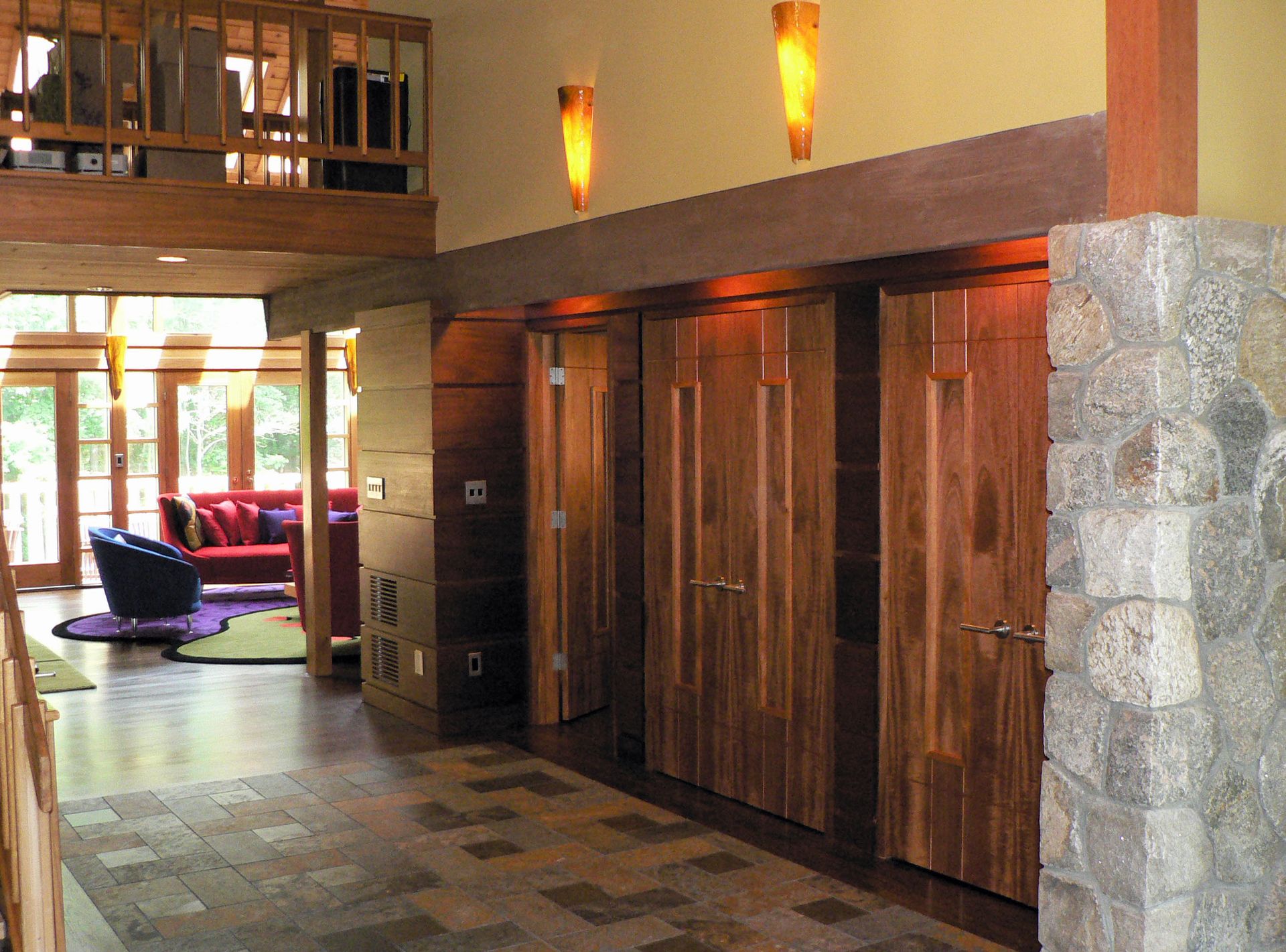 Hallway with wooden doors, stone column, and a loft. Sunlight streams through a doorway to a living room.