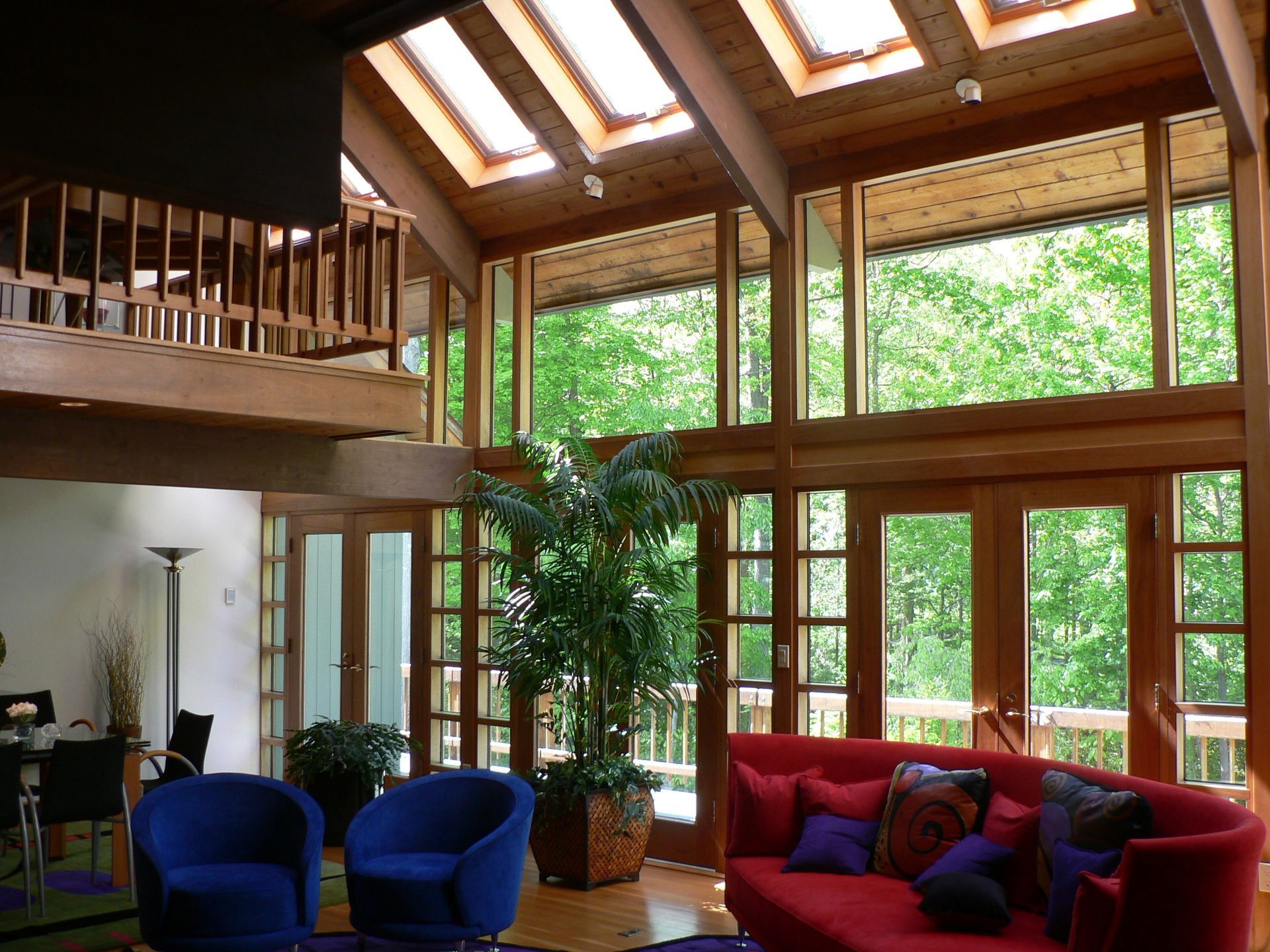 Living room with red sofa, blue chairs, large windows overlooking trees, and skylights.