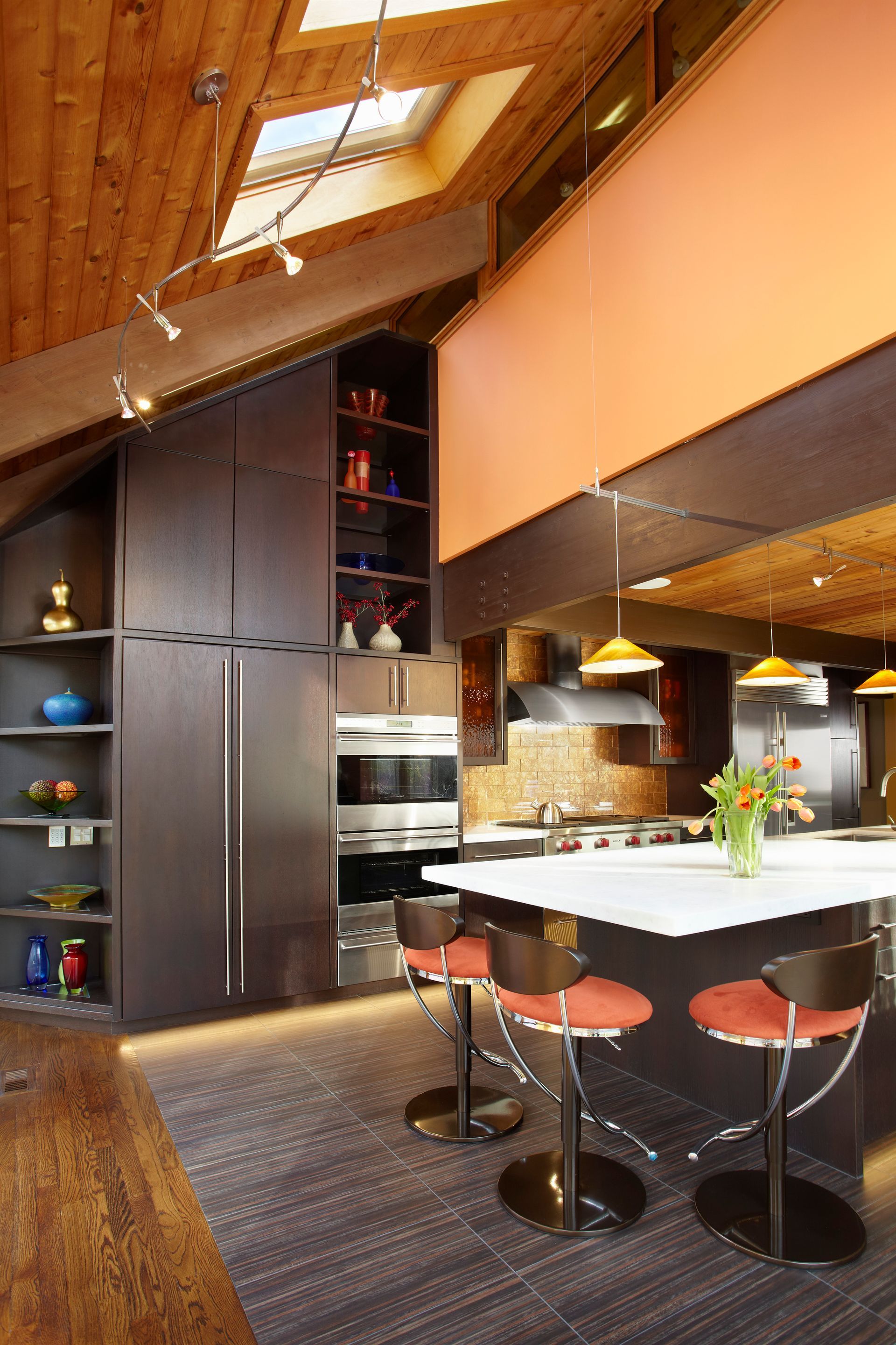 Modern kitchen with dark cabinetry, orange bar stools, island, and wood ceiling.