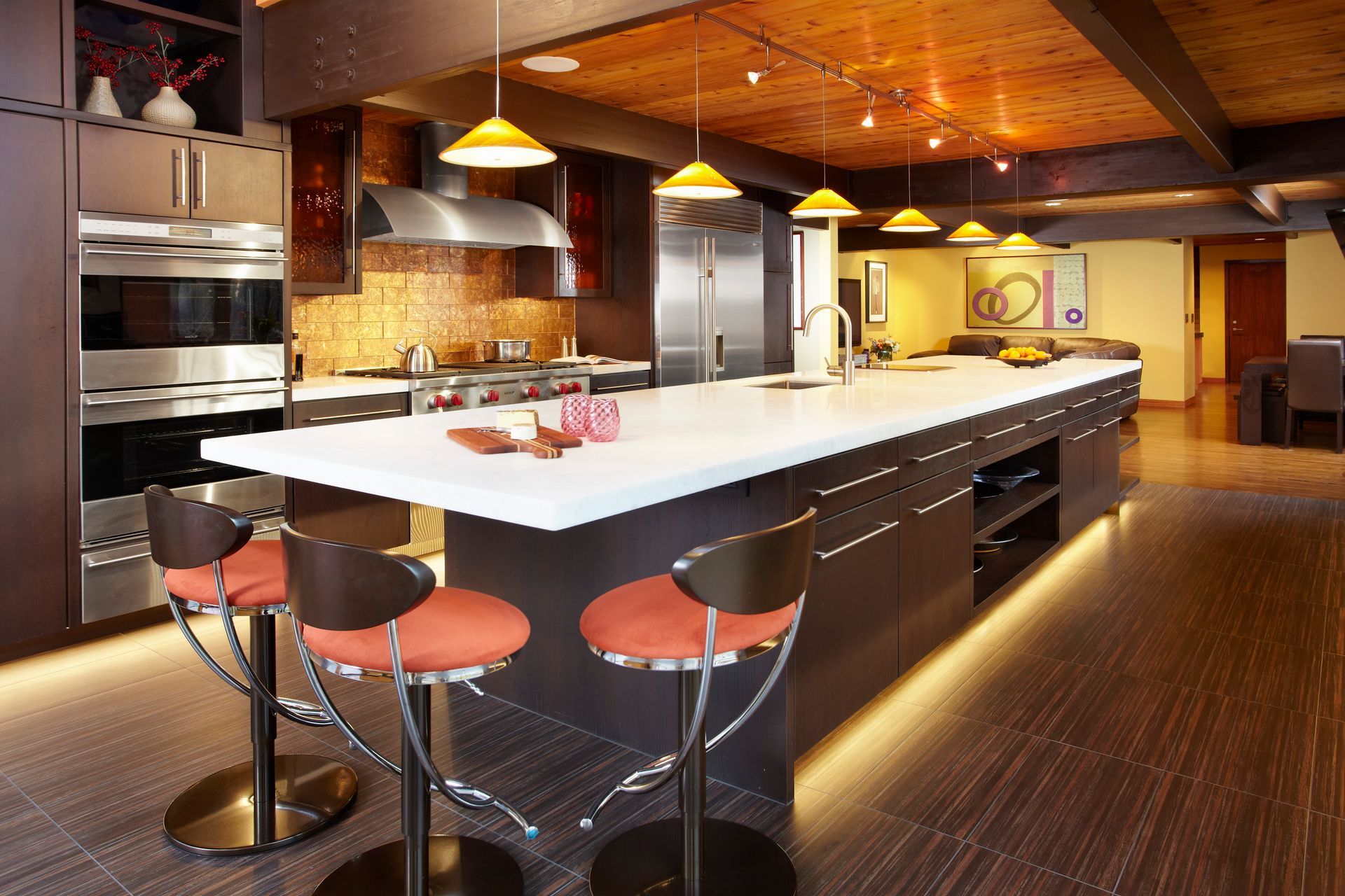 Modern kitchen with large island, stainless steel appliances, orange bar stools, and warm lighting.