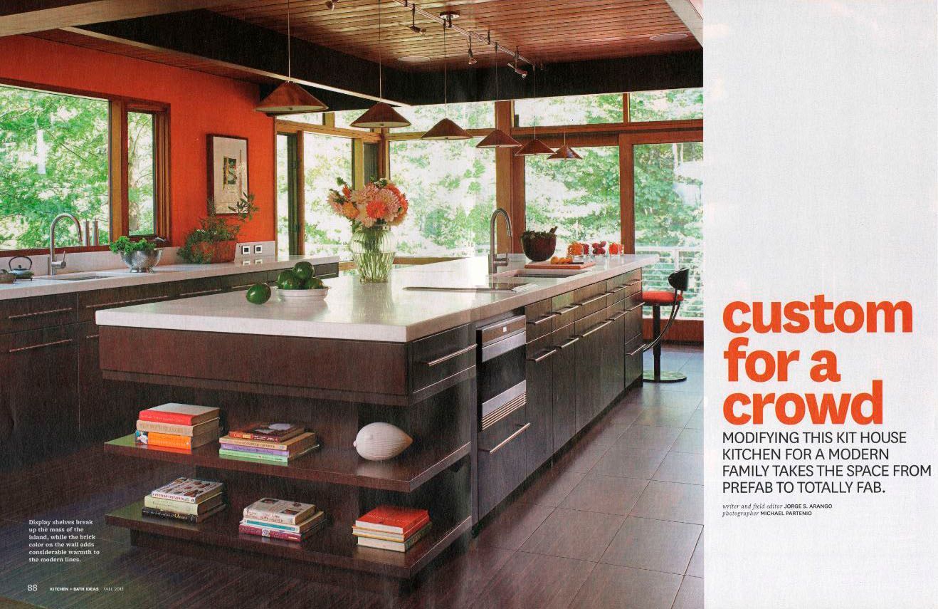 Modern kitchen with large island, wood cabinets, orange accent wall, and large windows with a lush green outside view.